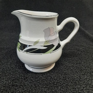May include: White ceramic creamer with a black and green floral design. The creamer has a rounded body and a handle.