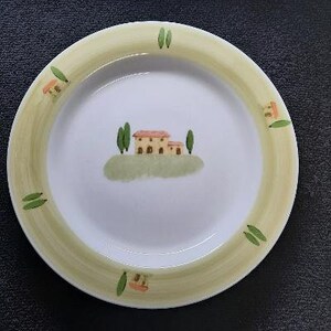 May include: A white ceramic plate with a green and yellow border. The plate features a painted scene of a small house with a red roof and green trees.