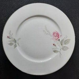 May include: A white ceramic dinner plate with a delicate floral design featuring a single pink rose and green leaves.