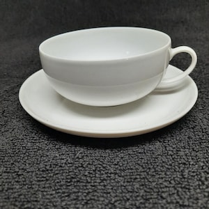 May include: A white teacup and saucer set. The cup has a rounded shape with a handle, and it sits on a matching saucer. The set is made of ceramic or porcelain and is suitable for serving tea or coffee.