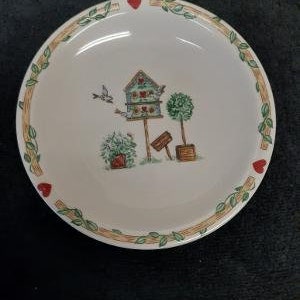May include: A white ceramic bowl with a green and brown leafy border and red hearts. The bowl features a hand-painted design of a birdhouse with a bird flying overhead, a bush, and a tree.
