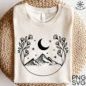 Può includere: Un'illustrazione in bianco e nero di una luna crescente, montagne e fiori in un cerchio. Il design è su una t-shirt color crema.