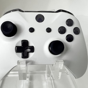 May include: A white Xbox controller with black buttons and joysticks. The controller is on a clear acrylic stand.