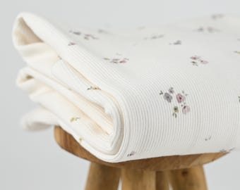 May include: Folded cream-colored blanket with a ribbed texture and a delicate floral pattern in shades of pink, gray, and green. The blanket is neatly stacked on a small wooden stool, showcasing its soft, inviting appearance.