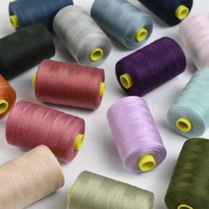 Könnte beinhalten: Eine Sammlung farbenfroher Garnrollen in verschiedenen Farbtönen, darunter Blau, Grün, Rosa und Lila. Jede Rolle hat einen gelben Kern. Die Fäden sind eng gewickelt, was darauf hindeutet, dass sie neu und für Näh- oder Bastelprojekte geeignet sind.