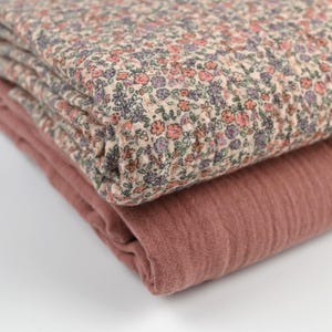 Könnte beinhalten: Zwei gefaltete Stoffe. Der obere Stoff hat ein Blumenmuster mit kleinen rosa, lila und grünen Blumen auf beigem Hintergrund. Der untere Stoff ist einfarbig, in einem gedämpften Rosaton. Die Stoffe sind übereinander gestapelt.