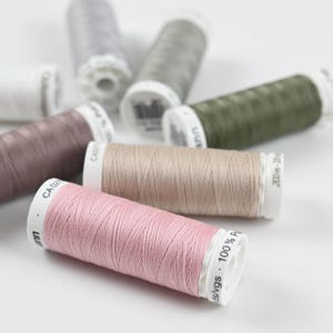 Könnte beinhalten: Eine Auswahl an Garnrollen in verschiedenen Farben, darunter Rosa, Beige und Grün. Die Rollen liegen auf einer weißen Oberfläche, einige sind scharf gestellt, andere unscharf. Die Rollen sind mit Text beschriftet.