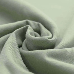 May include: Close-up of a soft, sage green fabric with visible texture and folds. The fabric appears to be a knit material, possibly cotton or a blend, with a subtle sheen. The image highlights the fabric's smooth surface and gentle curves.