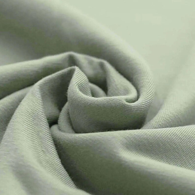 May include: Close-up of a soft, sage green fabric with visible texture and folds. The fabric appears to be a knit material, possibly cotton or a blend, with a subtle sheen. The image highlights the fabric's smooth surface and gentle curves.