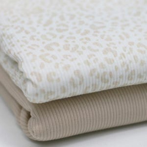 Op de afbeelding: Twee stukken geribbelde stof, een met een witte achtergrond en een beige luipaardprint, de andere effen beige.