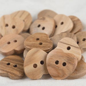 Könnte beinhalten: Eine Ansammlung von herzförmigen Holzknöpfen. Jeder Knopf hat zwei Löcher zum Annähen und zeigt ein natürliches Holzmaserungsmuster. Die Knöpfe variieren leicht in Farbe und Größe, wodurch eine rustikale Ästhetik entsteht. Ideal für Bastelprojekte.