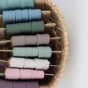 Può includere: Un cesto di vimini contiene bobine di nastro di tessuto colorato. I nastri sono in varie tonalità pastello, tra cui blu, verde, grigio e rosa. Ogni bobina è ben arrotolata e presentata su un bastoncino di legno. Il cesto è realizzato in materiale tessuto naturale.