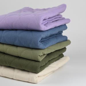 Könnte beinhalten: Ein Stapel gefalteter, gesteppter Stoffe in verschiedenen Farben: Lavendel, Blau, Olivgrün und Creme. Der Stoff hat ein rautenförmiges Muster. Das Bild zeigt eine Vielzahl von gefalteten Textilien.