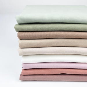 Könnte beinhalten: Ein Stapel gefalteter Stoffe in verschiedenen gedämpften Farben, darunter Salbeigrün, Braun und Rosa. Der Stoff scheint ein weiches Strickmaterial zu sein, und der Stapel ist ordentlich vor einem weißen Hintergrund angeordnet. Die Farben erzeugen eine beruhigende, neutrale Palette.