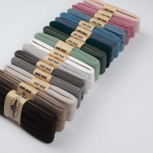 Könnte beinhalten: Eine Sammlung farbiger Stoffbänder, jedes mit einem Papierband mit der Aufschrift "oaki doki" umwickelt. Die Bänder sind diagonal angeordnet und zeigen eine Vielzahl von Farben, darunter Rosa, Blau, Grün, Grau und Braun. Die Bänder sind ordentlich gestapelt.