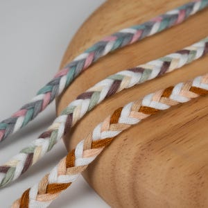 Può includere: Primo piano di tre cordoncini intrecciati in vari colori. I cordoncini sono realizzati in tessuto intrecciato e presentano un mix di colori tra cui grigio, rosa, bianco, marrone e pesca. I cordoncini sono disposti diagonalmente su una superficie di legno.