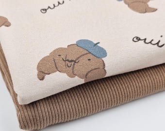 May include: Beige fabric with a brown and blue cartoon croissant design and the text "oui!" repeated. The fabric is folded over a brown ribbed fabric.