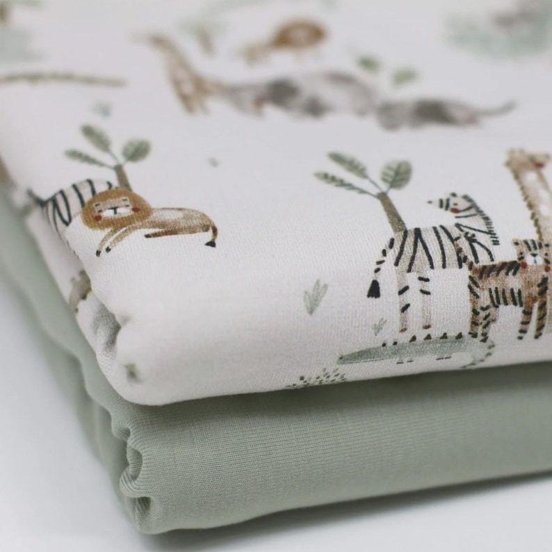 May include: Two stacks of fabric. The top fabric is white with a safari animal print featuring lions, giraffes, and zebras in brown and tan. The bottom fabric is a solid sage green color.