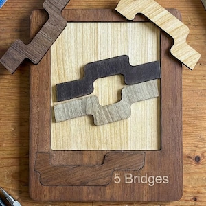 Puede incluir: Rompecabezas de madera con el texto "5 Bridges" y varias piezas. Un plano enrollado, lápices, una regla y gomas de borrar también están sobre la superficie de madera. El rompecabezas tiene un diseño de madera clara y oscura.