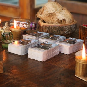 May include: A wooden table setting with lit candles, tarot cards, and decorative bottles. A wooden bowl holds a loaf of bread. The scene evokes a mystical or spiritual theme.