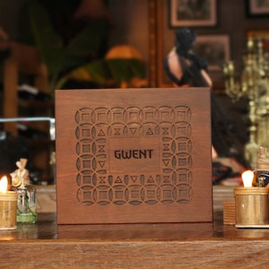 May include: A dark brown wooden box with the word "GWENT" carved into the front. The box has a decorative geometric pattern. Two bamboo candle holders with lit candles are in front of the box.