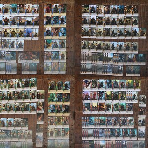 May include: A collection of trading cards featuring various characters and scenes, arranged on a wooden surface. The cards display detailed illustrations and are organized in multiple stacks and rows, showcasing a variety of fantasy-themed artwork.