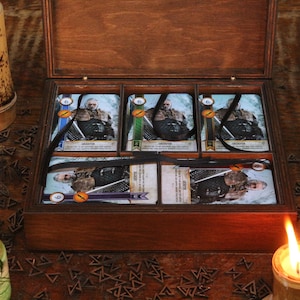 May include: A wooden box with a hinged lid, containing several game cards. The cards feature illustrations of a character and are tied with black ribbons. Decorative bottles and candles surround the box, creating a rustic aesthetic.