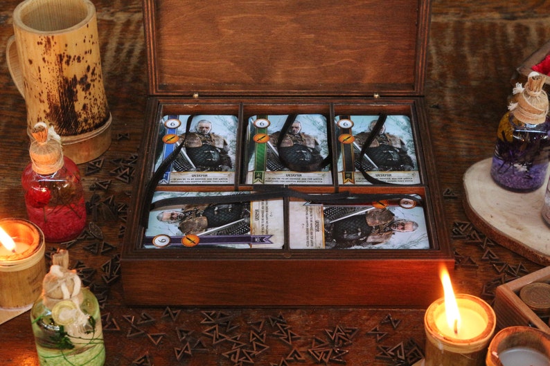 May include: A wooden box with a hinged lid, containing several game cards. The cards feature illustrations of a character and are tied with black ribbons. Decorative bottles and candles surround the box, creating a rustic aesthetic.