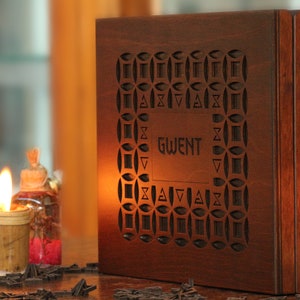 May include: A wooden box with the word "GWENT" carved into the front. The box has a decorative geometric pattern around the text. A lit candle and other decorative items are in the background.