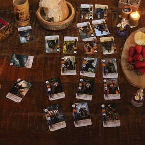 May include: A wooden table displays a board game with illustrated cards, food, and candles. The game cards feature fantasy characters. Fresh strawberries, green plums, and apple slices are on a wooden cutting board. A wooden box with a red interior is open.