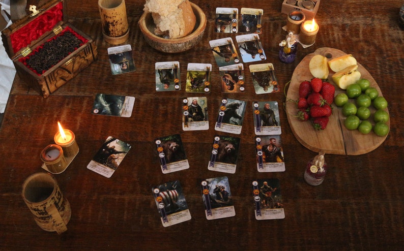 May include: A wooden table displays a board game with illustrated cards, food, and candles. The game cards feature fantasy characters. Fresh strawberries, green plums, and apple slices are on a wooden cutting board. A wooden box with a red interior is open.