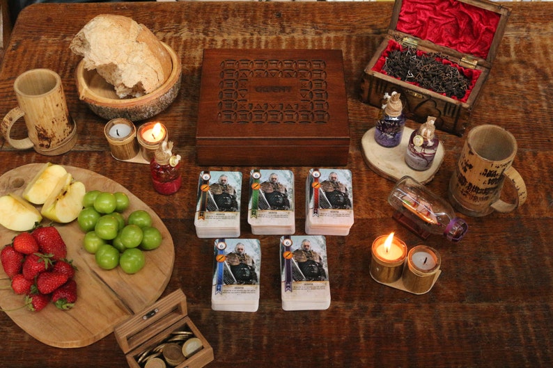 May include: A wooden table setting with a variety of items. Includes a wooden box, playing cards, fruit, bread, candles, and decorative bottles. The scene evokes a medieval or fantasy theme, suitable for a board game or themed event.