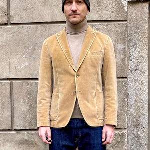 Può includere: Un blazer in velluto a coste beige con un maglione a collo alto marrone e jeans blu scuro. Il blazer ha una chiusura con un solo bottone e due tasche anteriori.