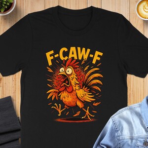 Könnte beinhalten: Schwarzes T-Shirt mit einem Cartoon-Hahn und dem Text "F-CAW-F". Der Hahn ist orange und rot mit überraschtem Gesichtsausdruck. Das T-Shirt wird auf einer Holzoberfläche präsentiert. Das T-Shirt ist kurzärmelig.
