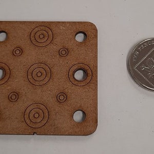 May include: A brown wooden square with multiple circles cut out. The circles are of varying sizes and are arranged in a pattern. The square has rounded corners and is about the size of a UK 20 pence coin.