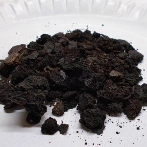 May include: A collection of dark brown, fragmented material, possibly soil or mulch, is arranged on a white, textured surface. The pieces are of varying sizes and textures, some coarse, others finer.