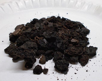 May include: A pile of dark brown, irregularly shaped pieces, possibly a type of soil or mulch, sits on a white, textured surface. The pieces vary in size and texture, with some appearing more coarse and others finer.