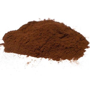 May include: A mound of finely ground, dark brown powder on a white backdrop. The powder has a textured appearance, with small particles visible. The overall shape is irregular, with a slightly rounded top and uneven edges.