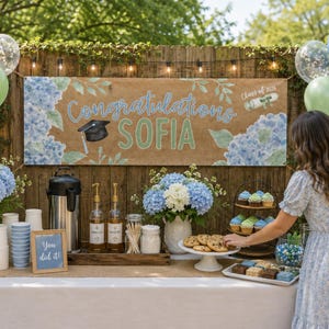 May include: A graduation party setup with a banner that reads "Congratulations SOFIA" with a mortarboard and scroll illustration. The banner is decorated with blue hydrangea flowers. The table has cupcakes, cookies, and drinks.