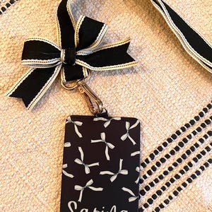 May include: Black and white patterned ID badge holder with a black and white ribbon lanyard and a gold clip. The badge holder has the name "Sarina" printed on it.