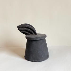 Może przedstawiać: Czarna rzeźba ceramiczna z cylindryczną podstawą i płaskim, okrągłym wierzchem. Zakrzywiony element w kształcie wachlarza wznosi się z góry. Rzeźba jest matowa czarna i stoi na białej powierzchni.
