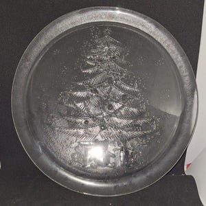 May include: A clear glass circular plate featuring an embossed design of a Christmas tree decorated with ornaments and a star on top. The base of the tree shows several wrapped gift boxes. The glass has a smooth rim and a textured center.