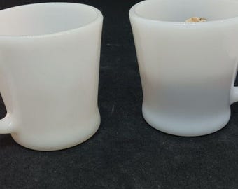 May include: Two white milk glass mugs with smooth, rounded bodies and C-shaped handles. The mugs feature a glossy finish and a simple, functional design with a slightly flared rim and a sturdy base, presenting a clean and uniform appearance.