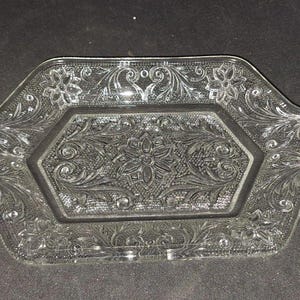May include: A clear pressed glass serving tray with a hexagonal center panel featuring a central floral motif surrounded by intricate scrollwork and leaf patterns. The tray includes two integrated side handles and a textured border design.