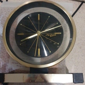 May include: A vintage City Quartz clock with a black face, gold hands, and a gold-colored frame. The clock has a rectangular base and a clear, circular outer casing. The text "City Quartz" is visible on the clock face.