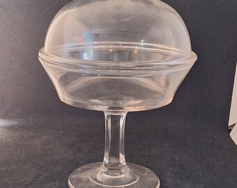 May include: A clear glass dome-shaped cake stand with a pedestal base. The stand has a knob on top of the dome.