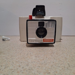 May include: A vintage Polaroid Swinger Land Camera Model 20, featuring a black and silver face with a prominent lens and flash. The camera has a red button and a black viewfinder. The camera is set against a cream-colored background.