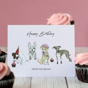May include: A white card with the text "Happy Birthday" and "From the Squad" with a cartoon illustration of four dogs wearing party hats and accessories. The card is surrounded by pink frosted cupcakes.