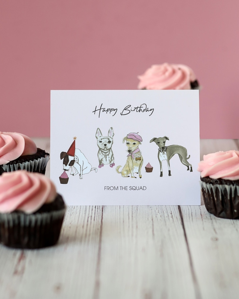 May include: A white card with the text "Happy Birthday" and "From the Squad" with a cartoon illustration of four dogs wearing party hats and accessories. The card is surrounded by pink frosted cupcakes.