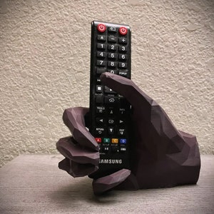 May include: A 3D printed hand holding a black Samsung TV remote control. The hand is dark brown and has a textured surface. The remote has a variety of buttons, including power, volume, channel, and menu buttons.
